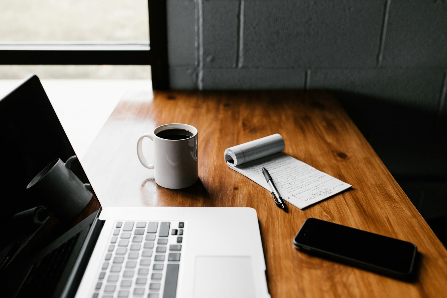 A laptop and coffee on a wooden desk in a bright workspace.